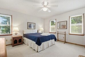 Une chambre à coucher comprenant un lit, deux tables de chevet avec des lampes, un ventilateur de plafond et de grandes fenêtres équipées de stores.