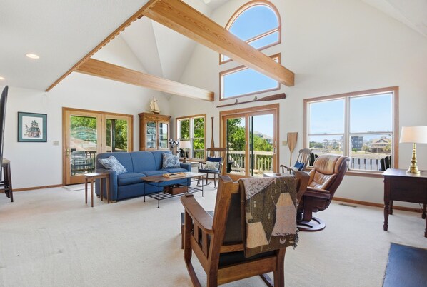 A spacious living room with a high ceiling, large windows, a blue sofa, a wooden chair, and a dining area.