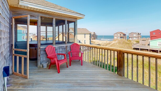 Een houten terras met twee rode stoelen en een glazen ruimte, met uitzicht op een strand en huizen op de achtergrond.