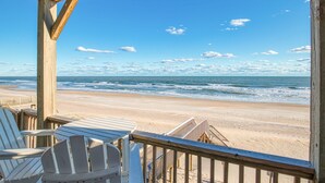Blick auf einen Strand von einer Holzterrasse mit weißen Stühlen, einem Holzgeländer und einem klaren, blauen Himmel.