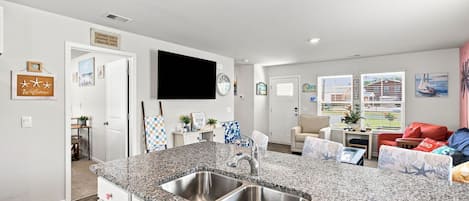 A modern kitchen with granite countertops and stainless steel sinks, a dining area with a table and chairs, and a living room with a sofa and a TV.