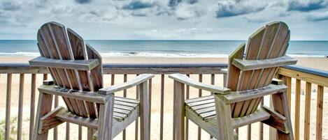 Two wooden Adirondack chairs facing a beach with a wooden deck and railing.