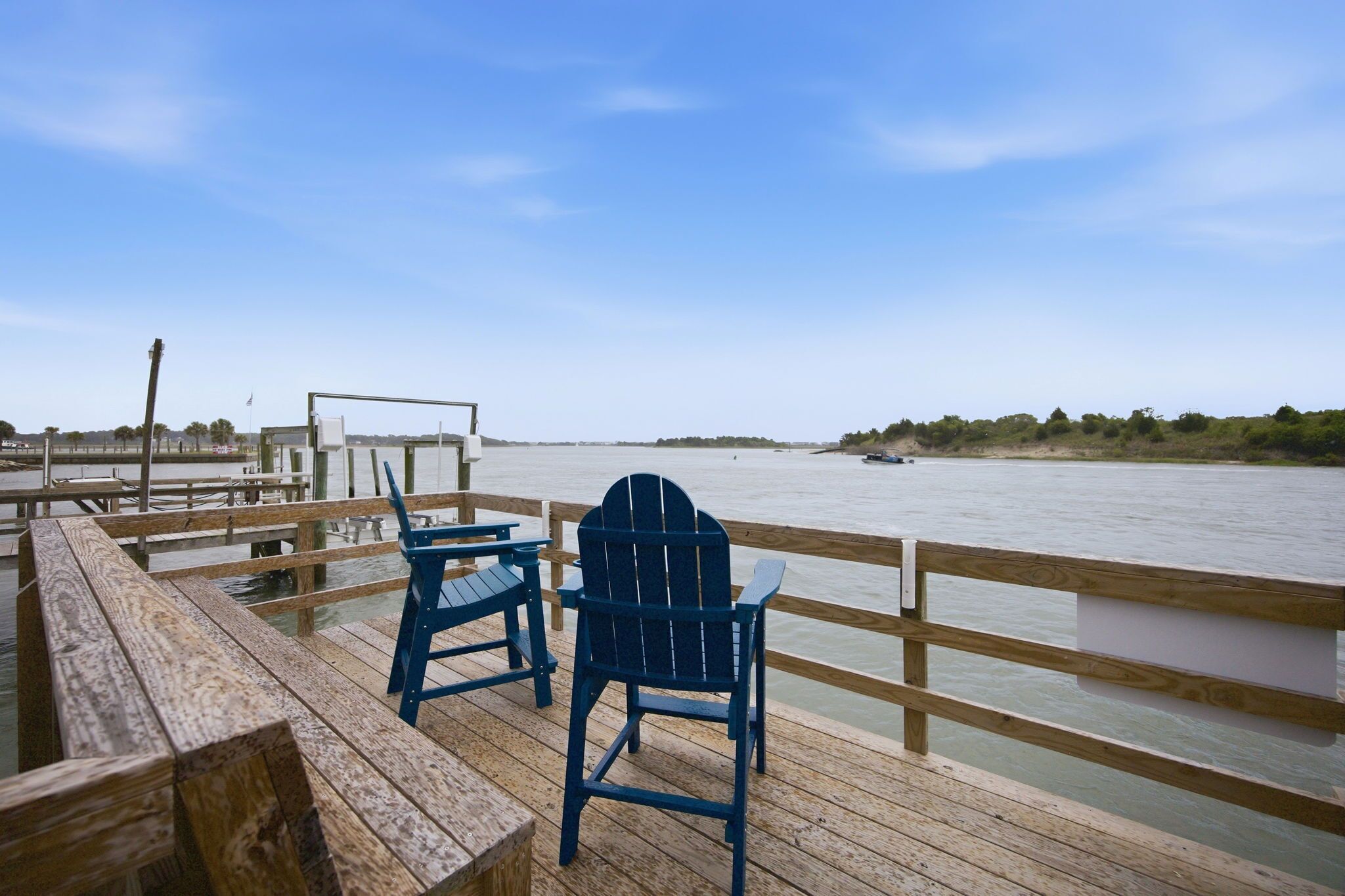 A wooden pier with two blue Adirondack chairs facing a calm river.