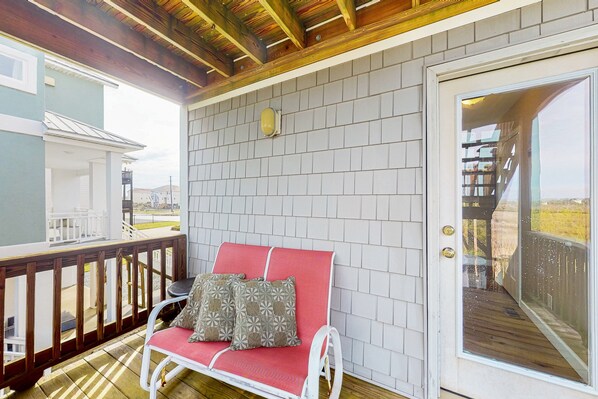 A covered porch with a red bench and patterned cushions, a wooden railing, and a view of the surrounding area.