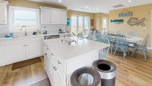 A kitchen with white cabinets, a large island, and a view of the ocean. There are blue chairs around a table, and a sign that says "topsail".