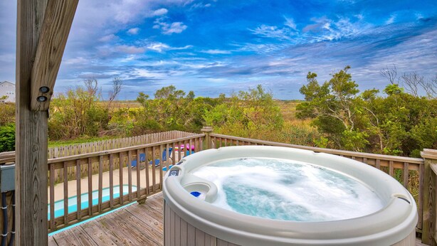 A hot tub on a wooden deck with a view of a swimming pool and trees.