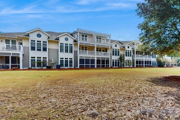 A multi-story residential building with a covered porch area, a grassy lawn, and a tree to the right.