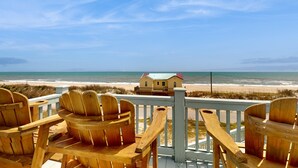 Una terraza de madera con sillas Adirondack con vistas a una playa y una casa con techo rojo.