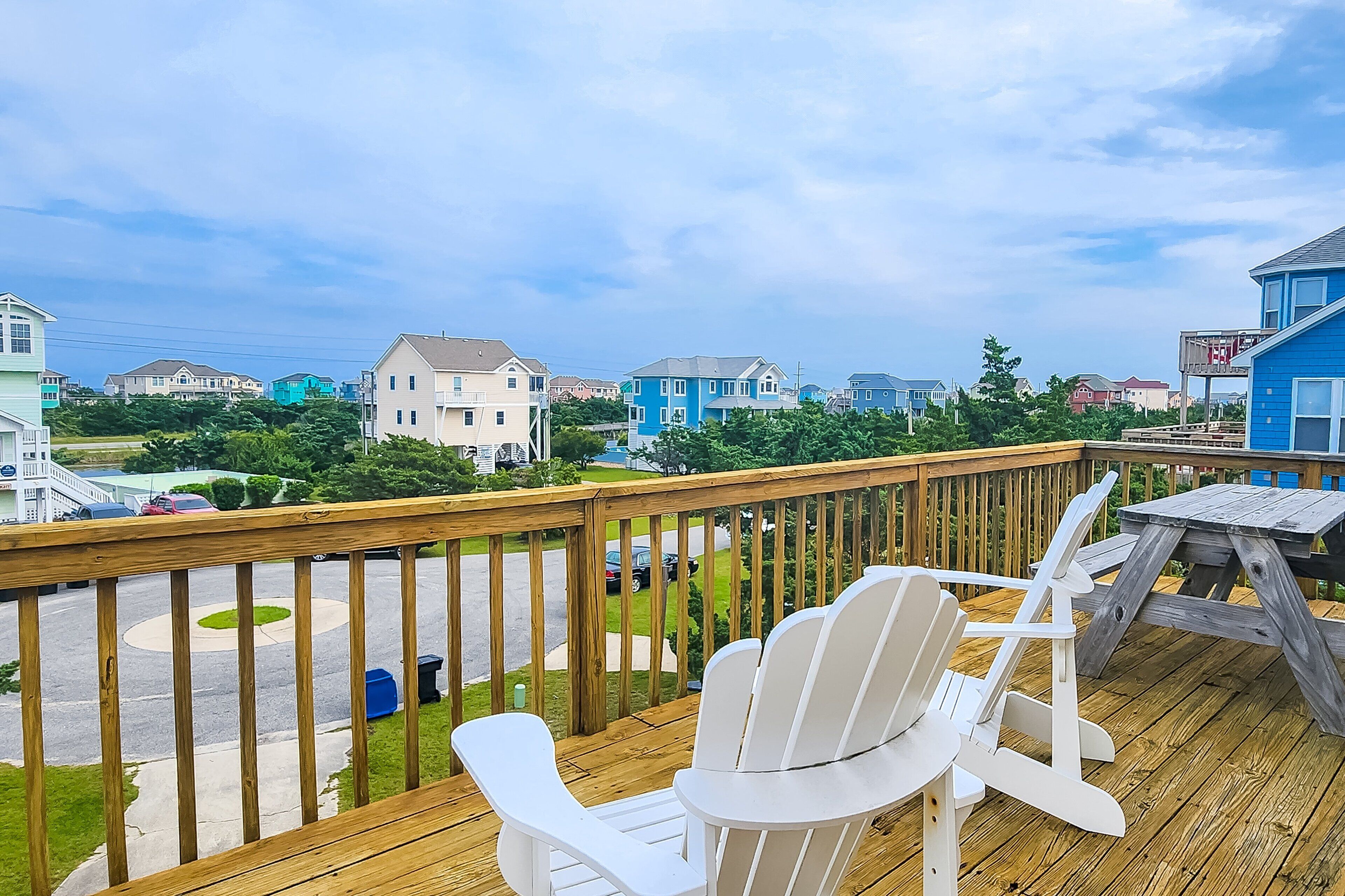 Une terrasse en bois, agrémentée de chaises Adirondack blanches et d’une table de pique-nique, qui offre une vue sur un quartier résidentiel avec ses maisons et sa route.