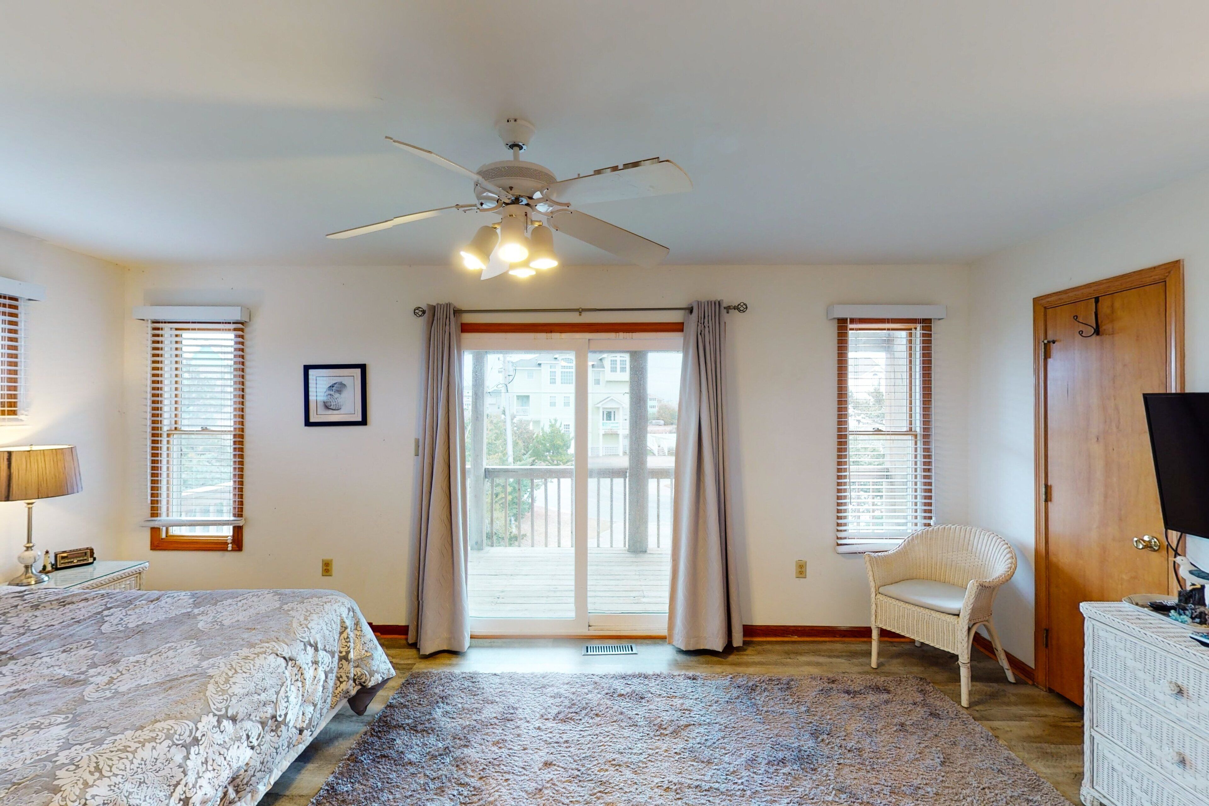 Une chambre à coucher comprenant un lit, un ventilateur de plafond, une chaise, une commode et une porte-fenêtre coulissante donnant sur l’extérieur.