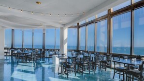 Un comedor amplio e interior con grandes ventanales que ofrecen vista al océano. Las mesas y sillas están dispuestas en filas.