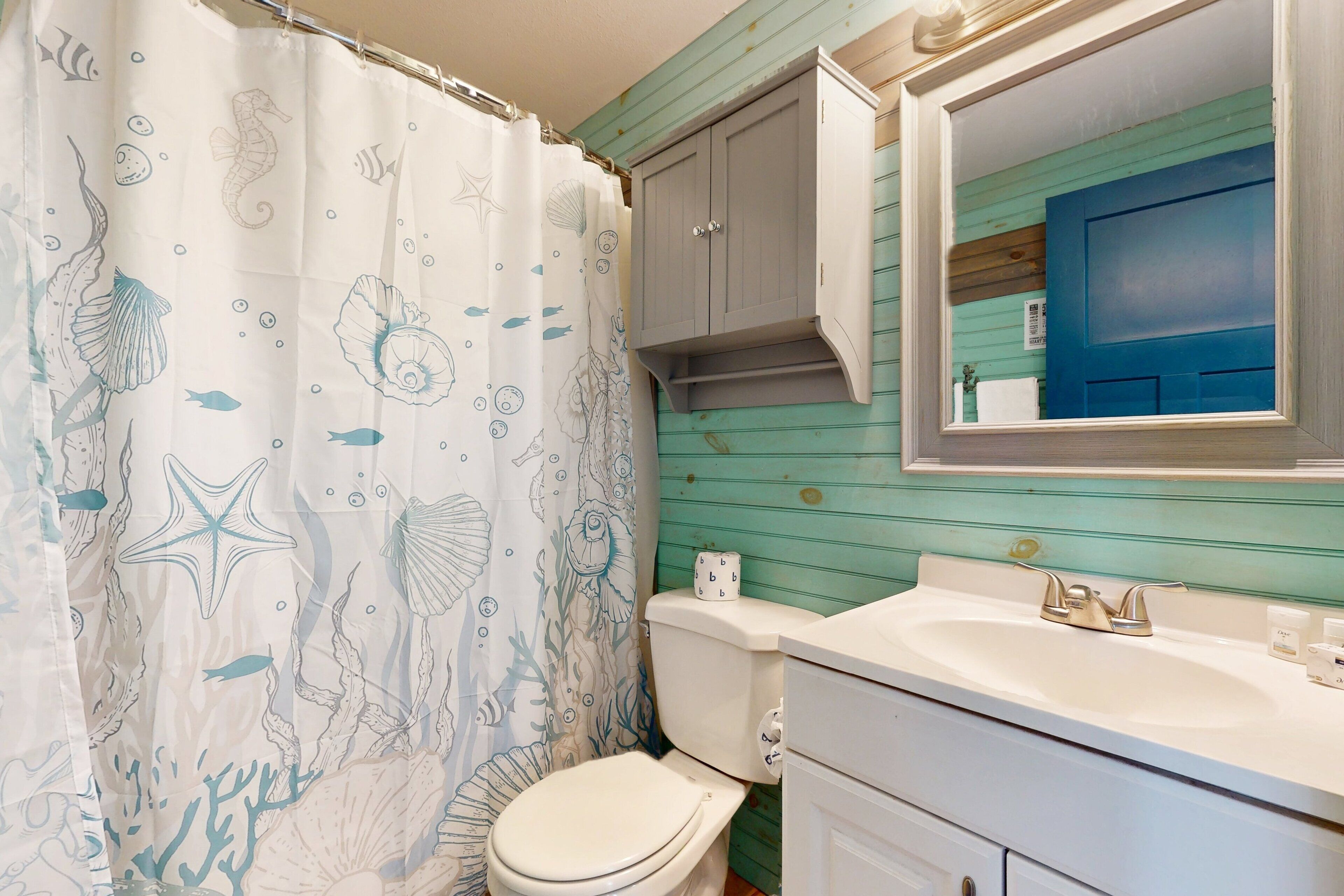 A bathroom with a seashore-themed shower curtain, a white toilet, a sink with a mirror above it, and a cabinet above the sink.