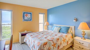A bedroom with a bed, two bedside tables, a nightstand, a chair, and a window with blinds. - Beachfront condo with private beach access, balcony, on-site pool, tennis & AC (North Topsail Beach)