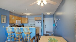 A kitchen with wooden cabinets, a microwave, a refrigerator, and a dining area with a table and chairs. - Beachfront condo with private beach access, balcony, on-site pool, tennis & AC (North Topsail Beach)
