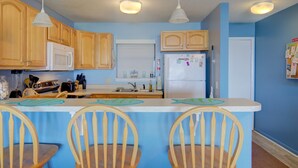 A kitchen with wooden cabinets, a white refrigerator, a microwave, and a sink. There are three wooden chairs at the kitchen island. - Beachfront condo with private beach access, balcony, on-site pool, tennis & AC (North Topsail Beach)