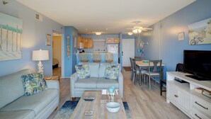 A living room with a sofa, coffee table, and TV. There is a dining area with chairs and a table. The walls are painted blue, and there are ceiling fans. - Beachfront condo with private beach access, balcony, on-site pool, tennis & AC (North Topsail Beach)