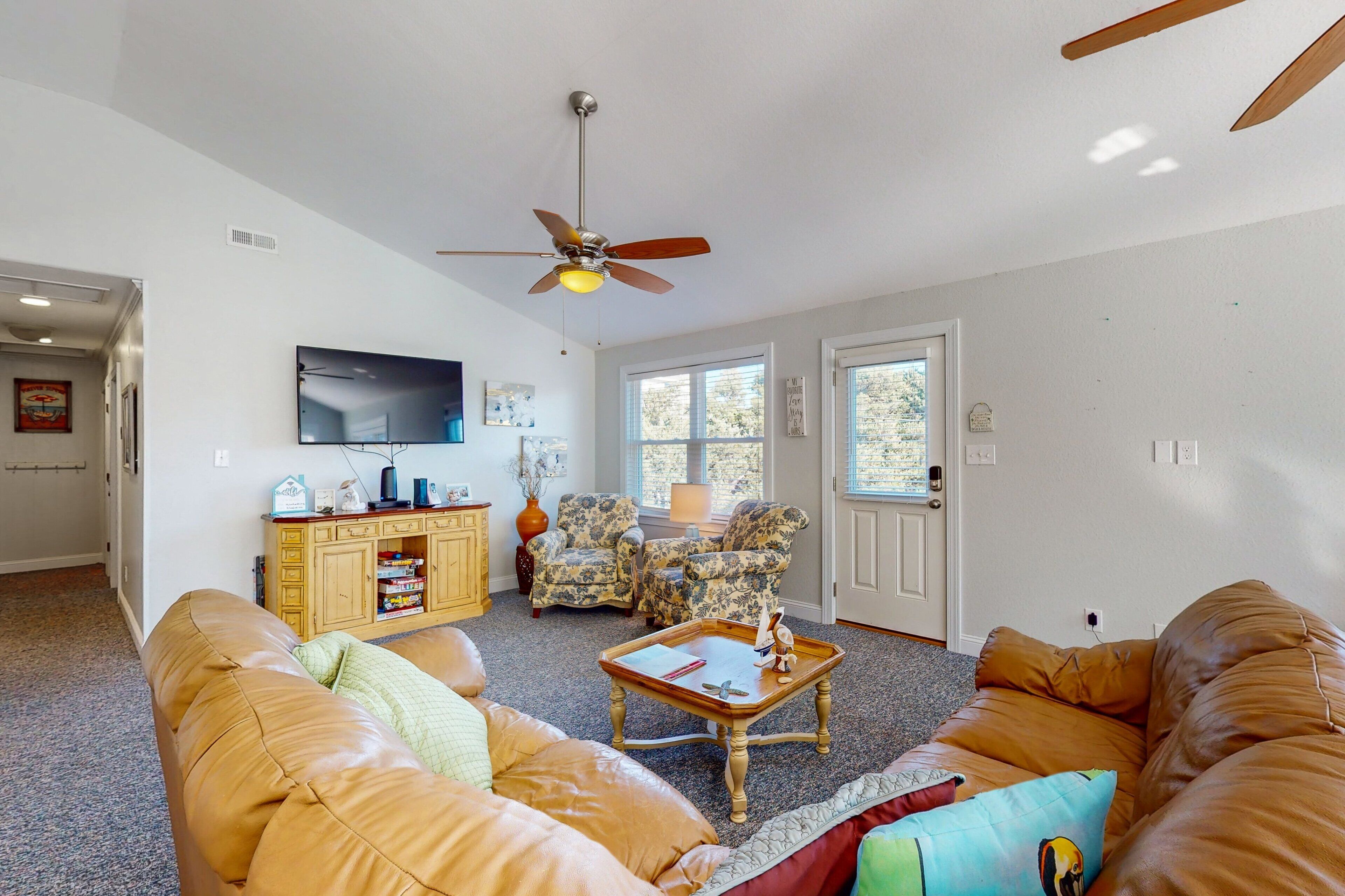 A living room with a brown leather couch, a wooden coffee table, and a ceiling fan. There is a television mounted on the wall and a door leading outside.