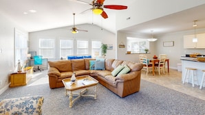 A spacious living room with a brown leather sectional sofa, a wooden coffee table, and a ceiling fan. There is a dining area with a table and chairs, and a kitchen area with a countertop and stools.