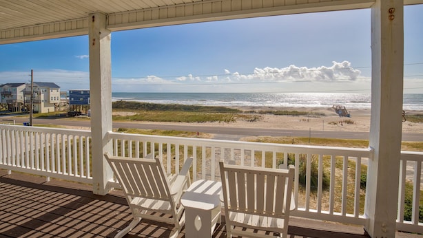 Une véranda couverte, dotée de balustrades blanches et de deux fauteuils Adirondack blancs, donnant sur une plage avec des vagues et une route en arrière-plan.