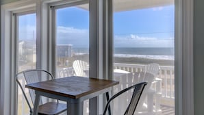 Une pièce avec une table et des chaises, une vue sur l’océan par de grandes fenêtres et un balcon avec des chaises blanches.