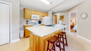 A modern kitchen with wooden cabinets, a white island with bar stools, a microwave, and a refrigerator. - Coastal condo with golf course views, on-site pools, gym, & balcony (Calabash)