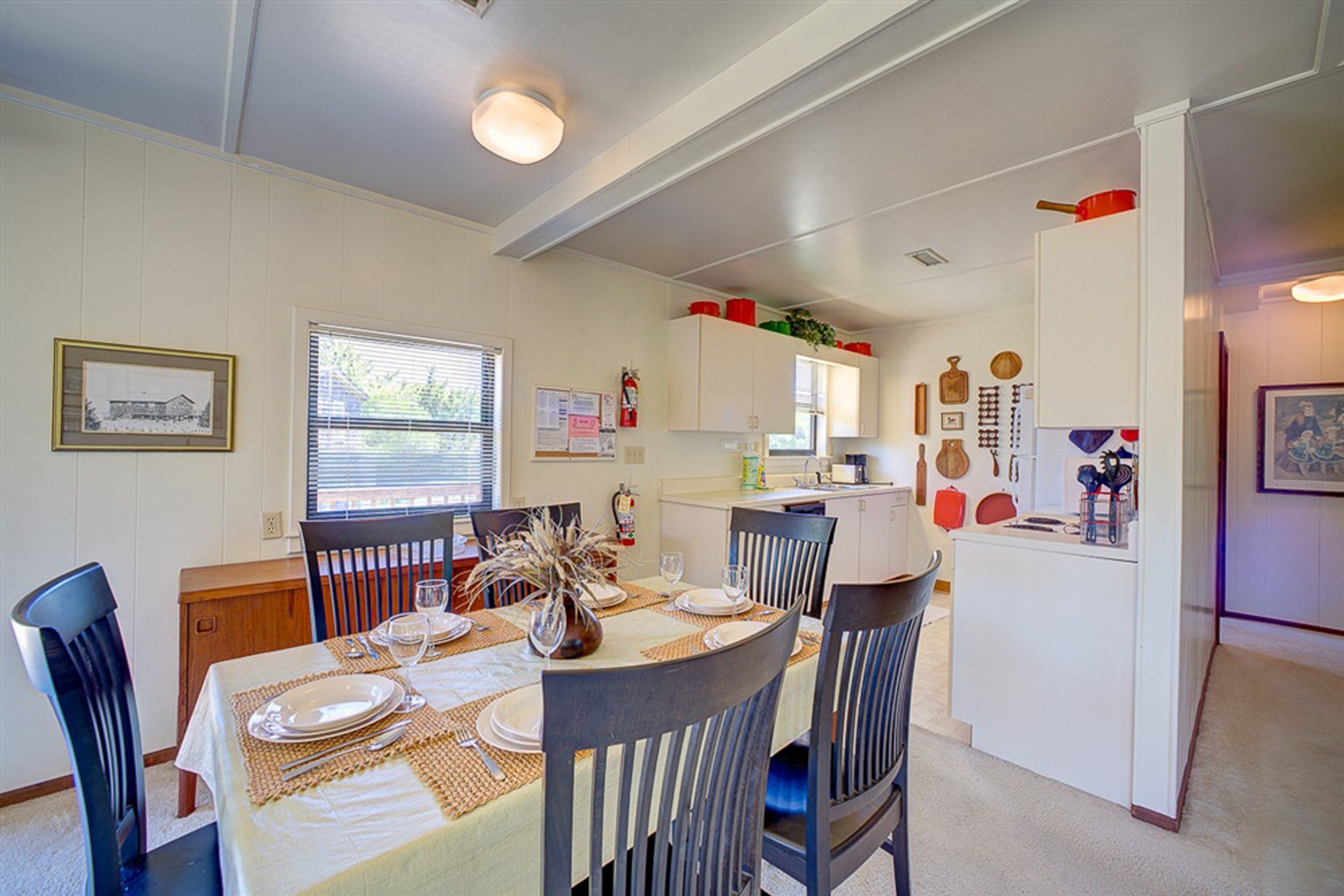Comedor con una mesa dispuesta para cuatro personas, sillas, una cocina al fondo y un refrigerador con adornos.