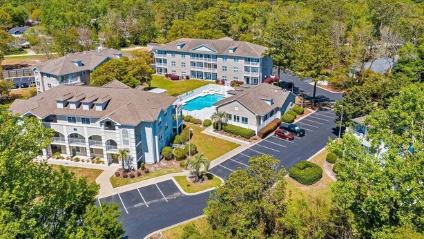 A multi-story hotel with a swimming pool, parking lot, and surrounding greenery. - Colony II - Unit 509 at Oyster Bay condo | fast WiFi, seasonal pools, hot tub, fitness room (Sunset Beach)