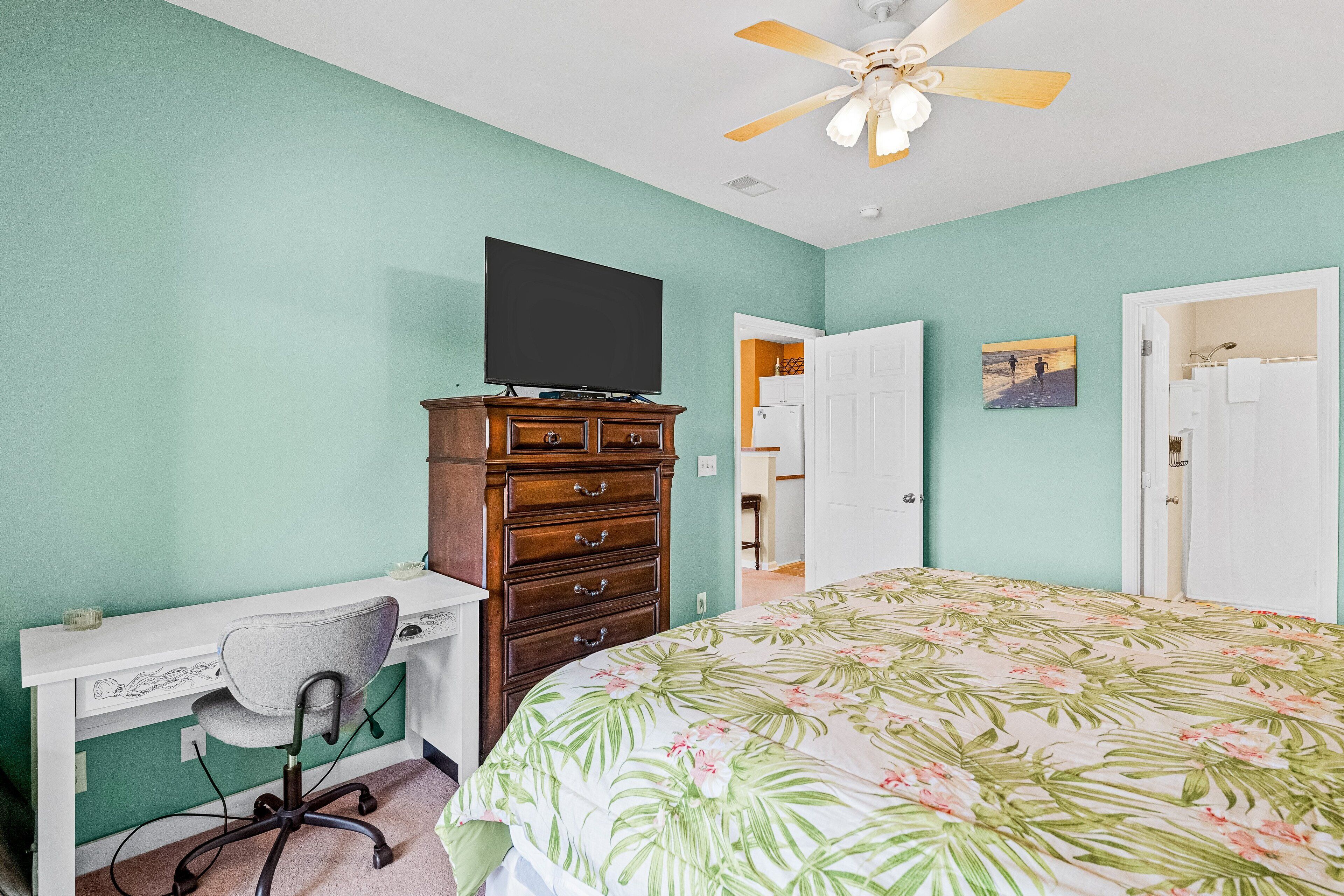 Een slaapkamer met een bed, een bureau met stoel, een dressoir, een plafondventilator en een deur naar een andere kamer.