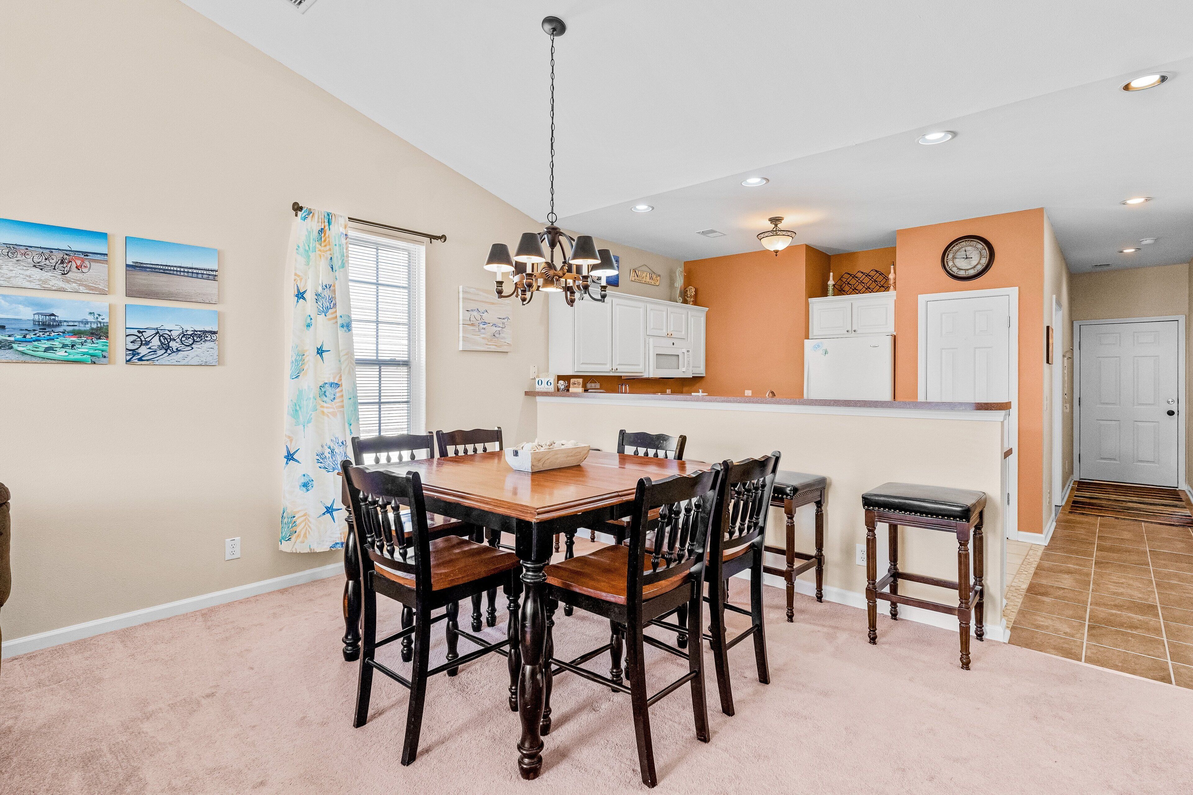 A dining area with a wooden table and chairs, a chandelier, and a kitchen in the background.