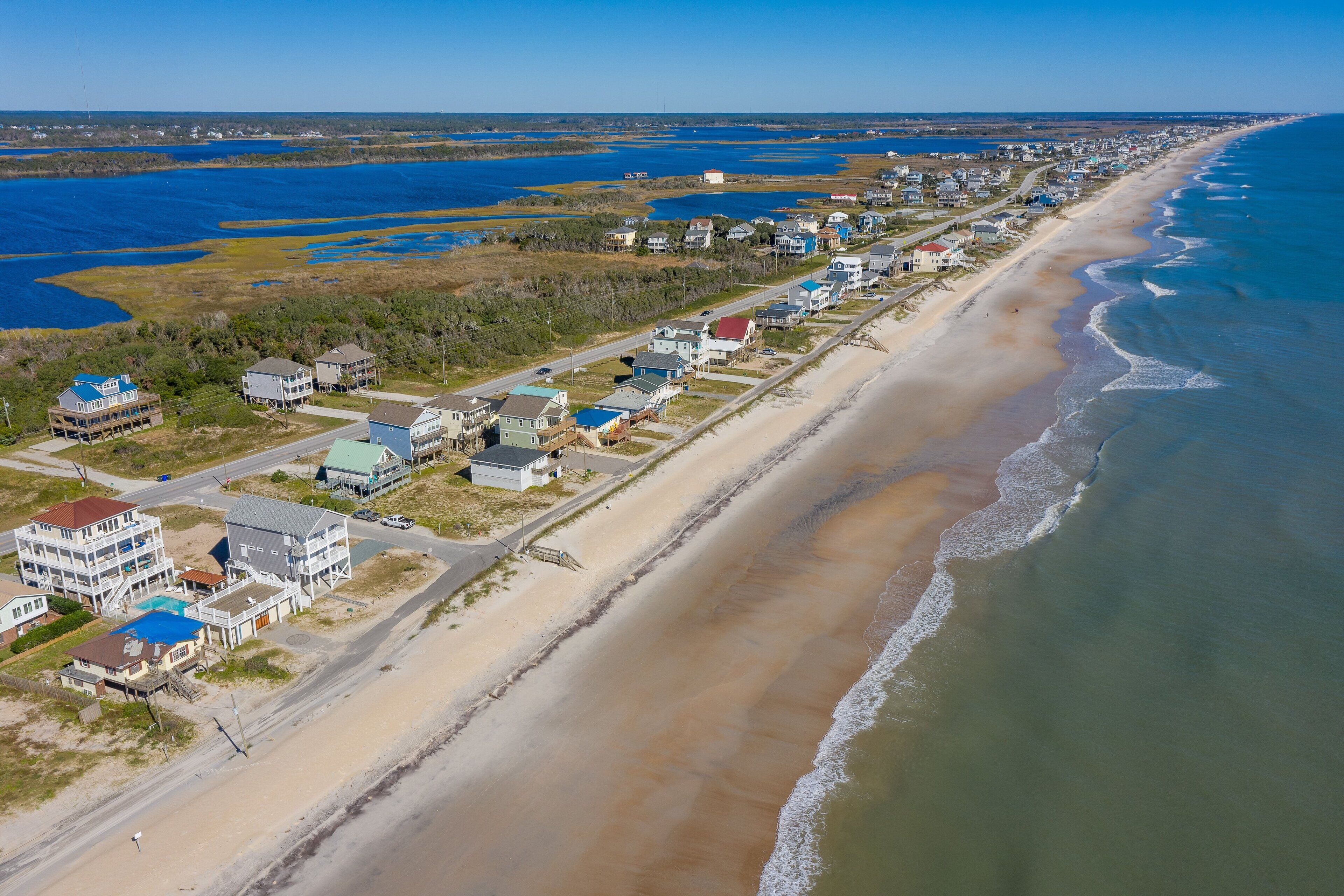 A coastal residential area with houses along the beach, a road, and a large body of water.