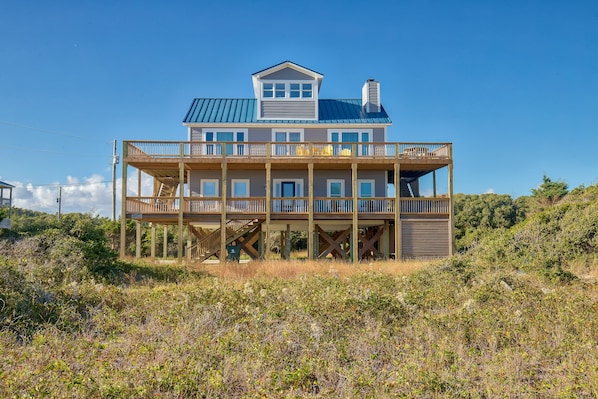 A two-story beachfront house with a balcony, a covered porch, and a staircase leading to the upper level.