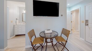 Ein Zimmer mit einem runden Glastisch, zwei Korbstühlen, einem flachbildfernseher an der Wand, einer weißen Kommode und einer Tür zu einem anderen Raum.