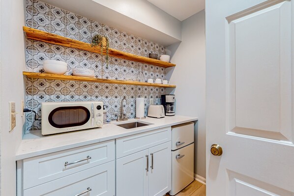 A kitchen with white cabinets, a microwave, a coffee maker, and a patterned backsplash. - Ev's Escape,201B: Lovely stay with patio, & kitchenette - dog- & snowbird-friendly (Sunset Beach)