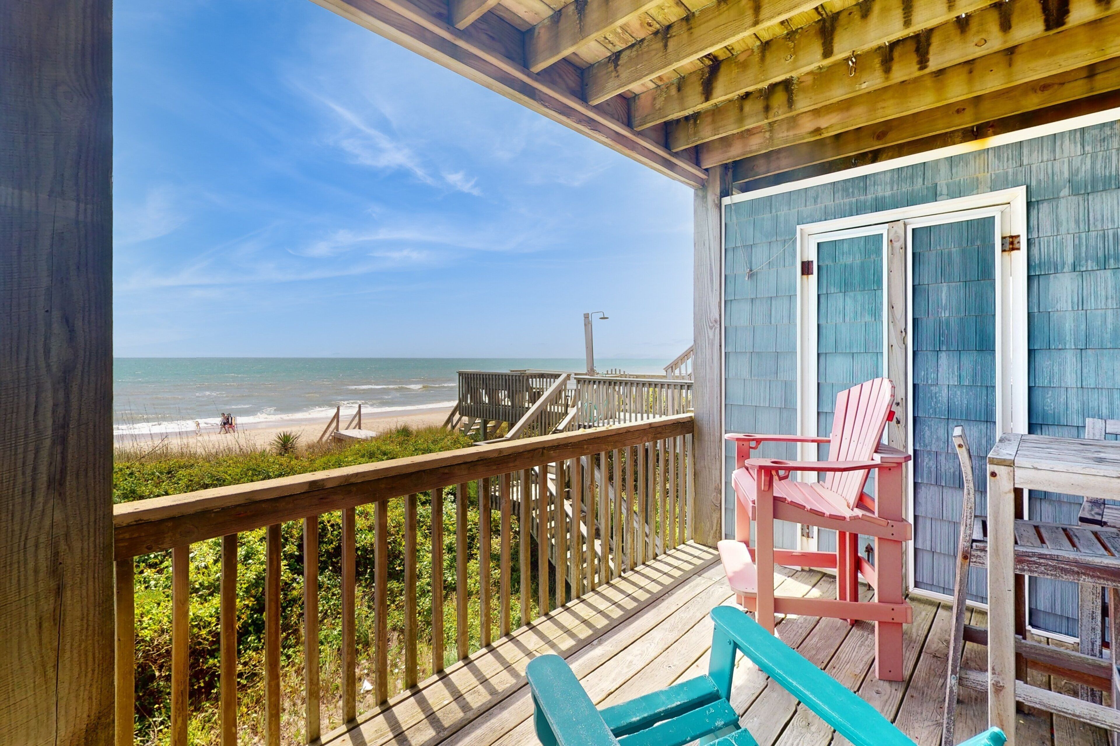 A wooden deck with a view of the beach, a blue building with a door, and two pink chairs.