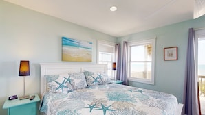 A bedroom with a bed, a nightstand, a lamp, a painting, and a window with curtains. - Oceanfront duplex with 4 decks, private beach walkway, central AC, & W/D (North Topsail Beach)