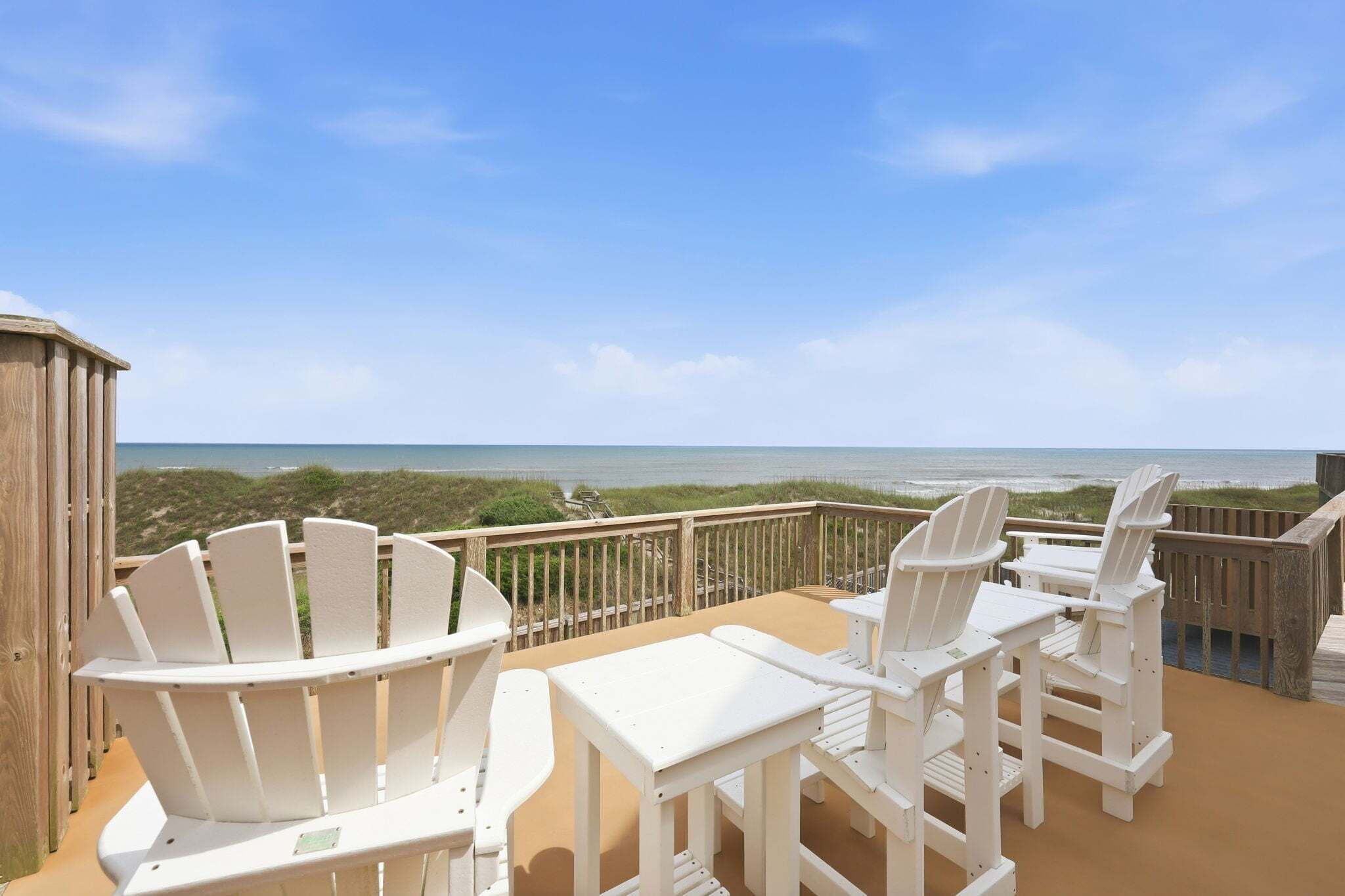 A beachside deck with white Adirondack chairs and a table, overlooking the ocean.