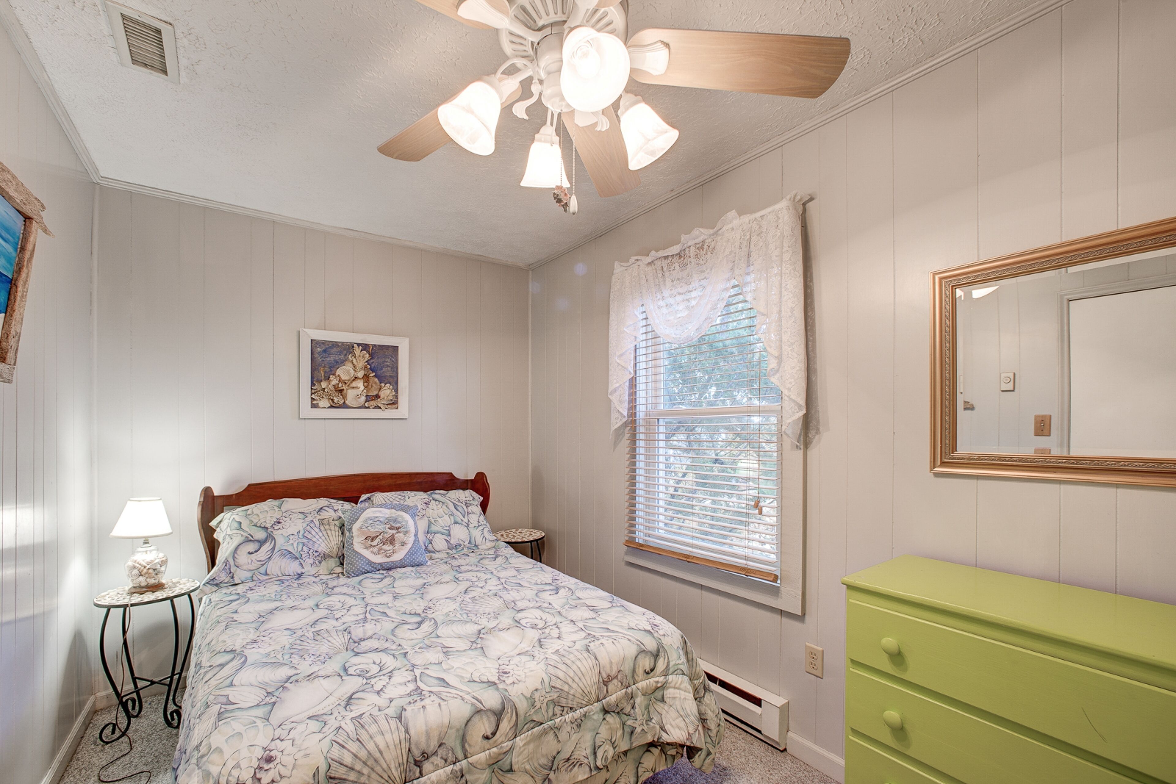 Une chambre à coucher avec un lit, une commode verte, un ventilateur de plafond, une fenêtre avec des rideaux et un tableau encadré accroché au mur.