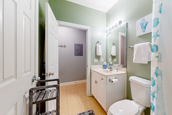 A bathroom with a toilet, sink, and mirror. There is a towel rack and a shower curtain with a sea shell pattern. - SB72B: Coastal Suite with golf course view and shared pool access (Sunset Beach)