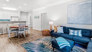 A modern living room with a blue sofa, a dining table with chairs, and a kitchen area in the background. - Crow Creek Retreat: Golf-course-front villa at Crow Creek with screened porch and washer/dryer (Calabash)