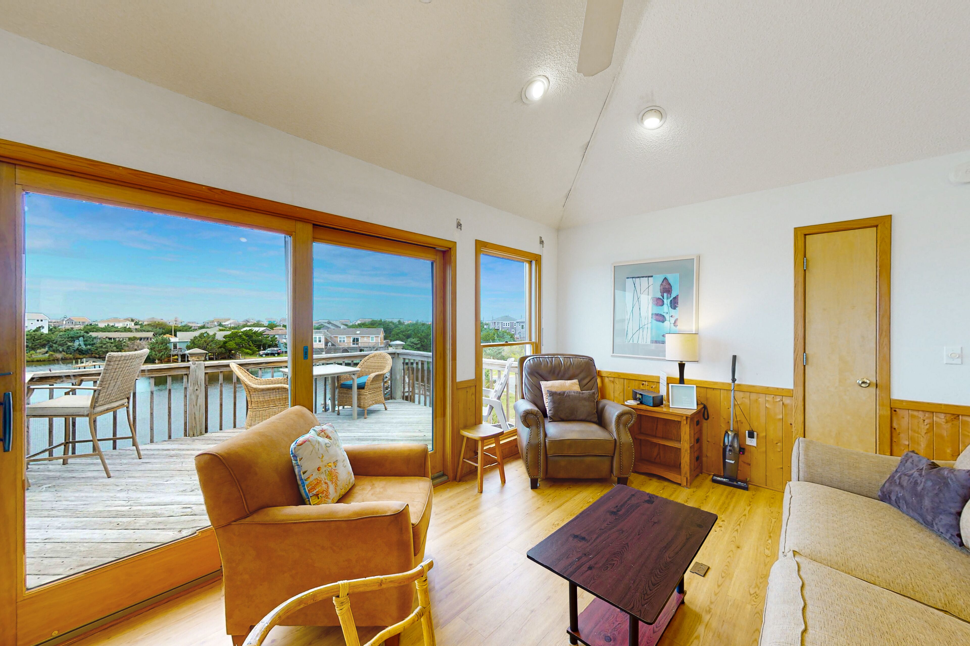 Een ruime woonkamer met een houten vloer, grote ramen, een bank, een fauteuil, een salontafel en uitzicht op een balkon met tuinmeubilair.
