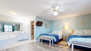 Une chambre bien éclairée, avec deux lits, une coiffeuse avec un miroir, un ventilateur de plafond et un téléviseur fixé au mur.
