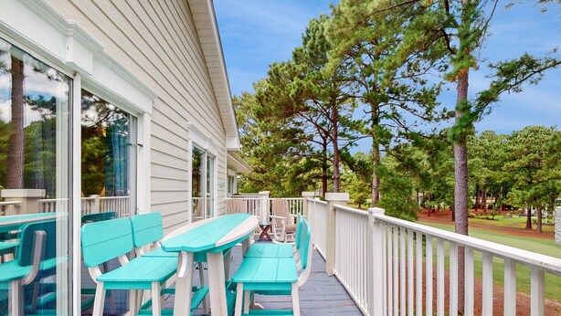 Un espace extérieur abrité, aménagé avec des chaises et des tables turquoise, des rampes blanches, et offrant une vue sur des arbres et une pelouse.