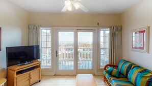 A living room with a television on a wicker stand, a colorful sofa, and a bench. - Oceanfront, dog-friendly condo with ocean view, central AC & WiFi (Hatteras)