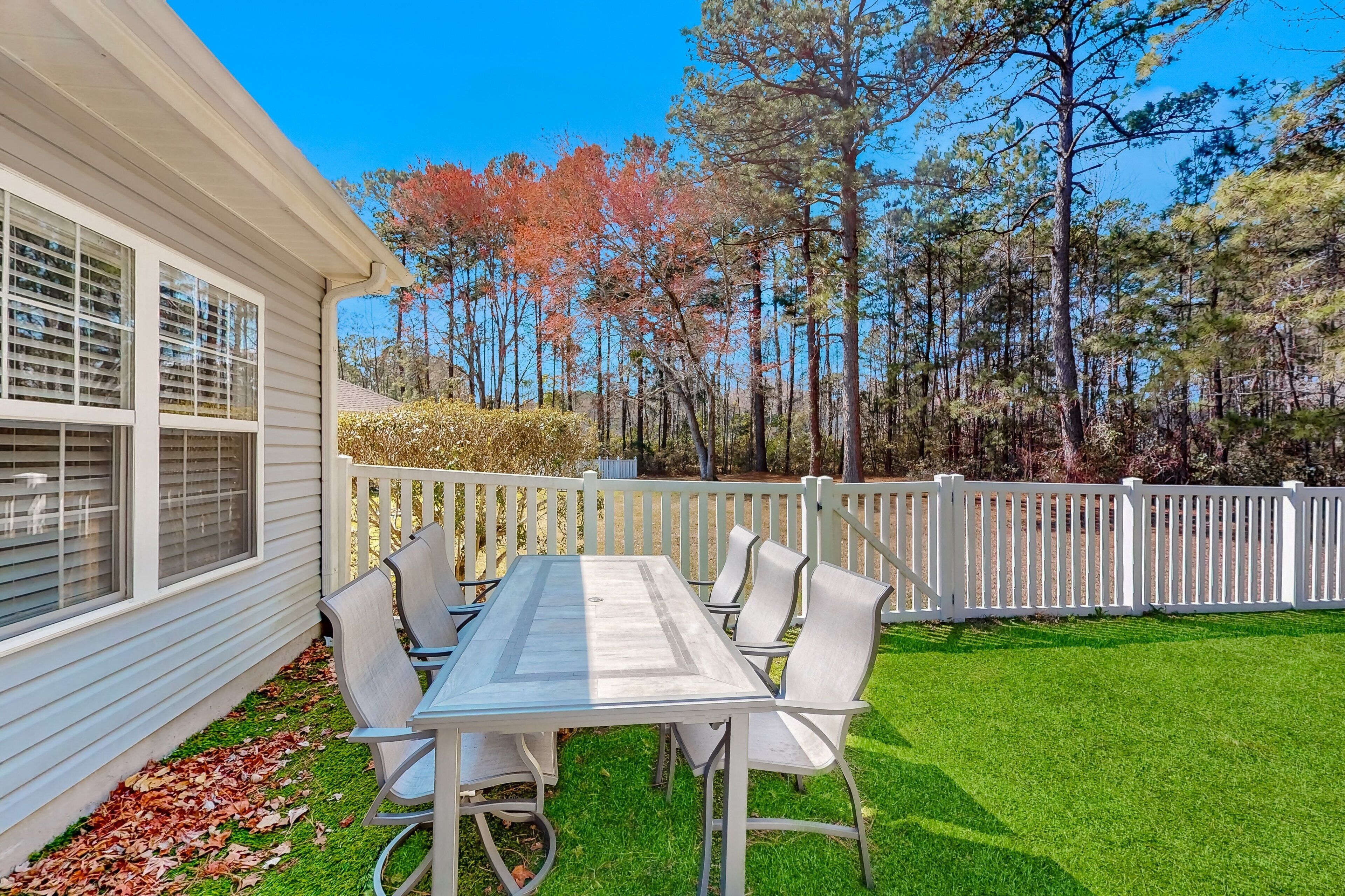 A patio with a table and chairs, a white fence, and trees in the background.