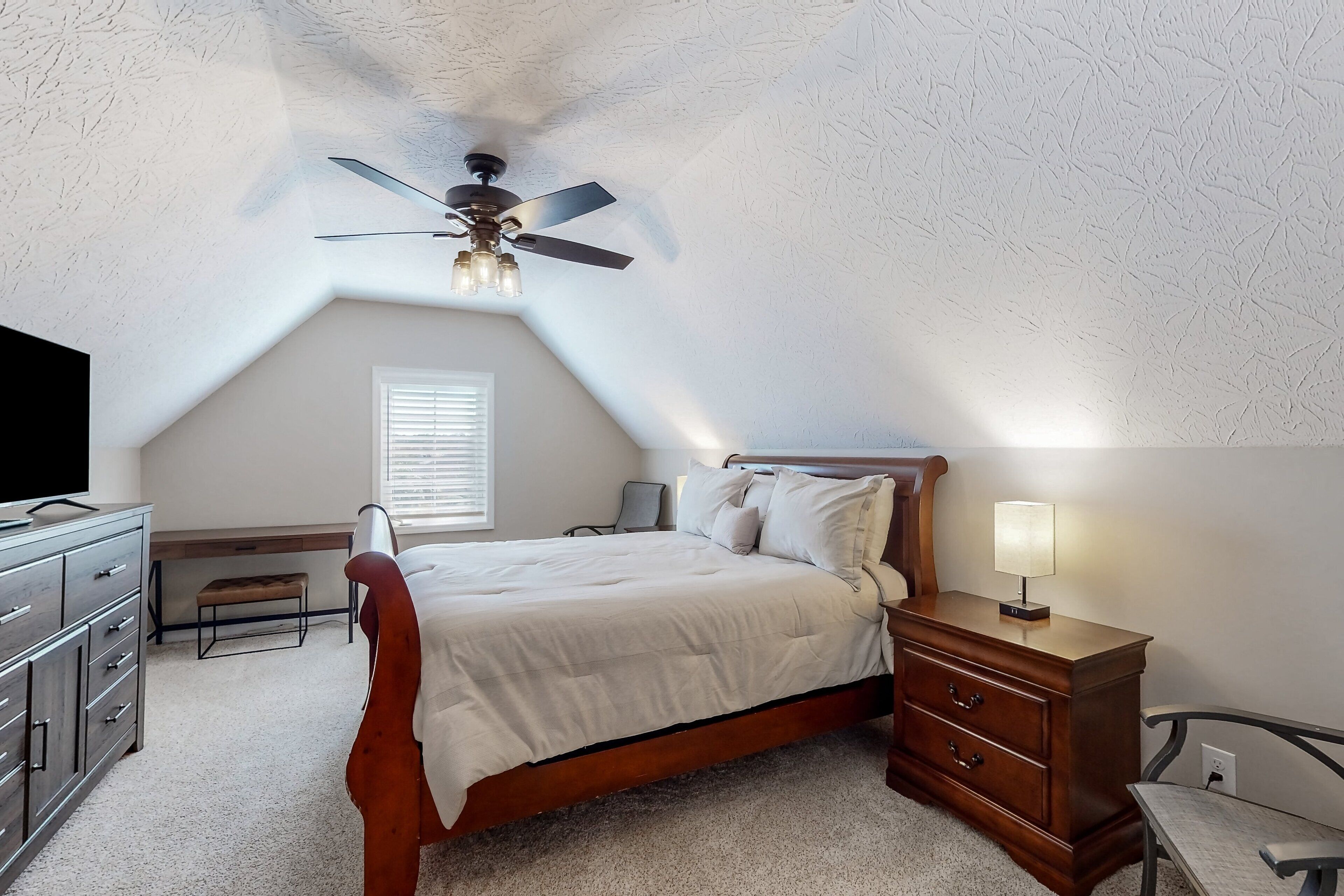 A bedroom with a sloped ceiling, a bed, a nightstand, a dresser, and a ceiling fan.