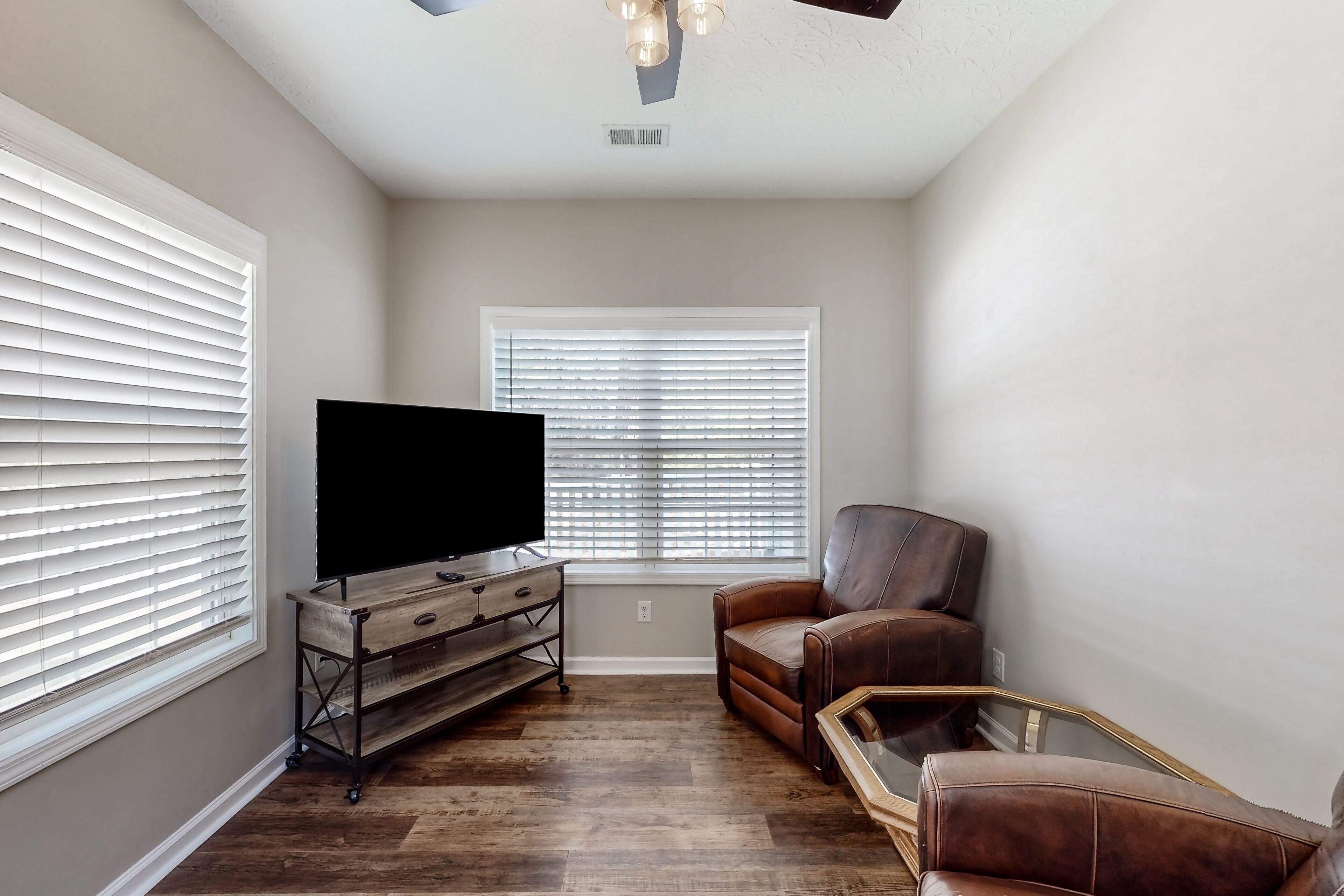 Un salon avec un téléviseur à écran plat posé sur un meuble en bois, deux fauteuils en cuir, une table basse en verre et une fenêtre avec des stores.