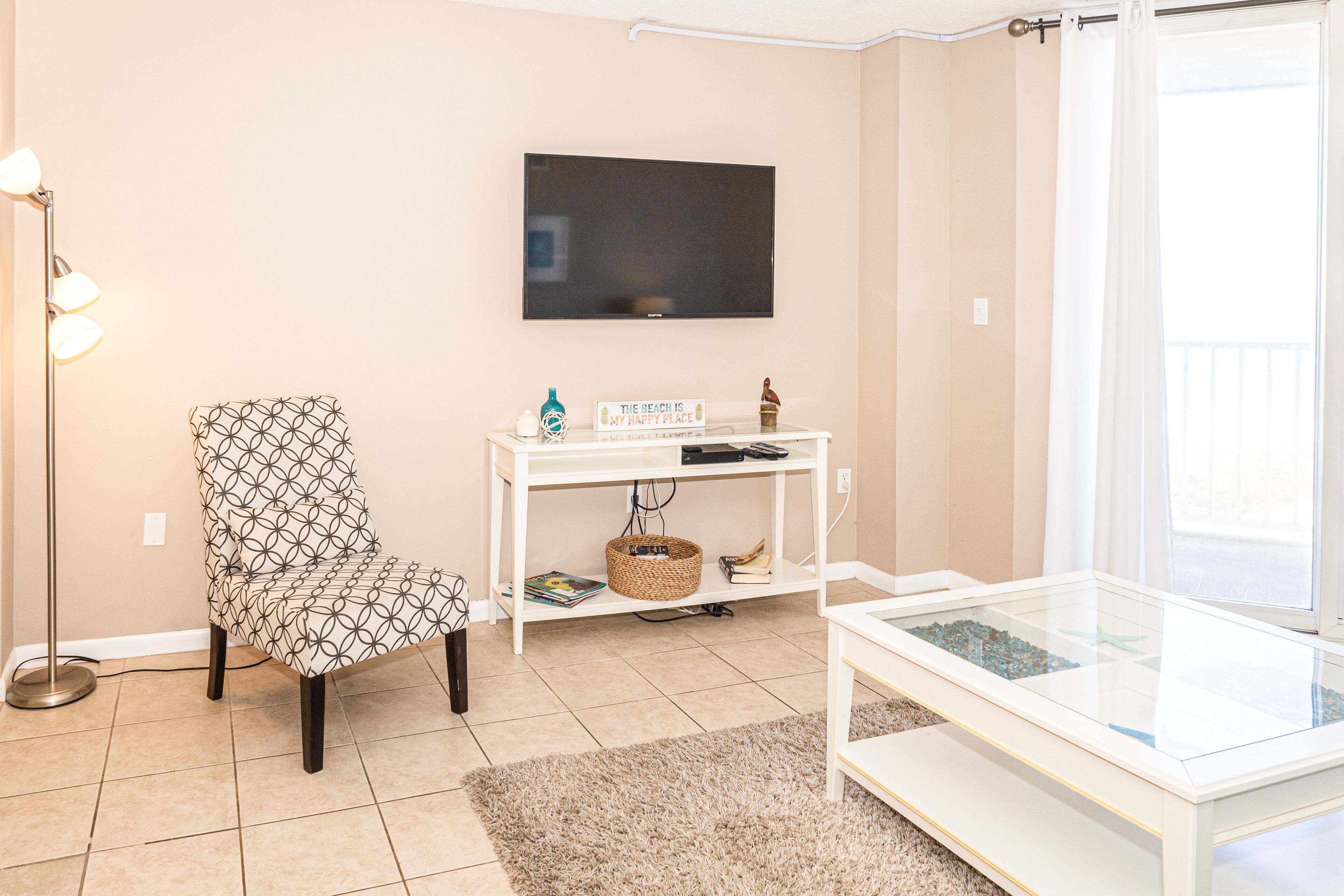 Un salon avec un fauteuil à motifs, une table d’appoint blanche sur laquelle est posée une télévision à écran plat, une table basse en verre et une vue sur l’extérieur par une fenêtre.