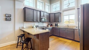 A modern kitchen with dark cabinets, a large island, and a view of the outdoors through large windows. - Slowin' Dune | Lavish Island Getaway with Golf Cart Near Beach, Golf, & Dining (Caswell Beach)