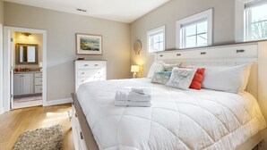 A bedroom with a large bed, a ceiling fan, a nightstand with a lamp, and a window with a view of trees. - Slowin' Dune | Lavish Island Getaway with Golf Cart Near Beach, Golf, & Dining (Caswell Beach)