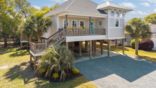 A two-story house with a covered porch, a deck, and a small garden area. - Slowin' Dune | Lavish Island Getaway with Golf Cart Near Beach, Golf, & Dining (Caswell Beach)